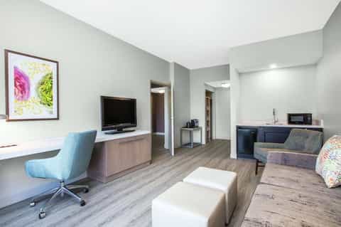 Modern hotel suite with desk, TV, seating area, and kitchenette with neutral tones
