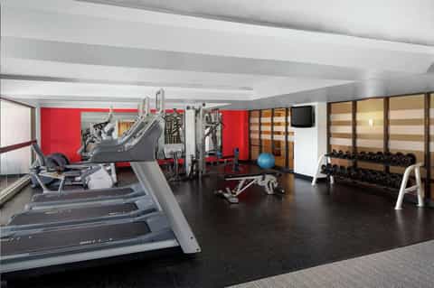Modern fitness center with treadmills, red accent wall, weight rack, exercise equipment, and dumbbell storage