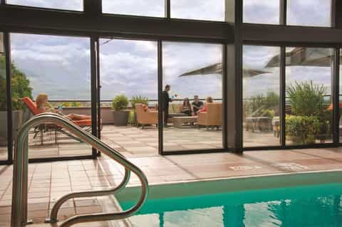 Indoor pool with metal handrail and floor-to-ceiling windows overlooking a rooftop terrace with guests