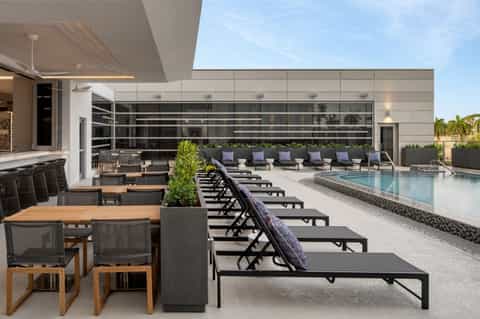 Modern rooftop pool deck with loungers, glass building facade, and palm trees under blue sky