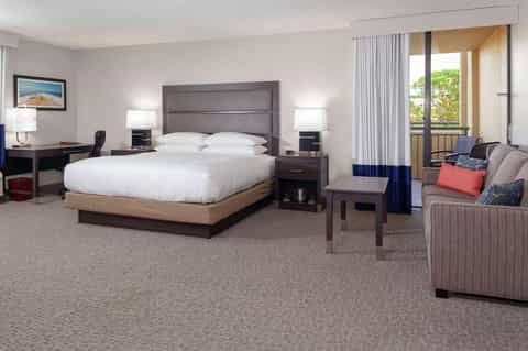 Spacious hotel suite with king bed, work desk, seating area, and balcony overlooking greenery