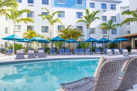 Resort pool surrounded by blue umbrellas, palm trees, and white multi-story building