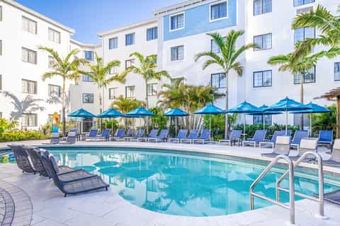 Resort pool surrounded by white buildings, blue umbrellas and palm trees under clear sky