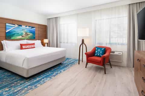 Modern hotel bedroom with white bedding, red accent chair, ocean view, and wood accents
