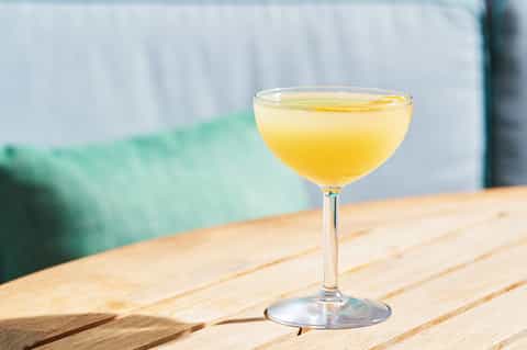 Yellow cocktail in a coupe glass on a wooden table with blurred green cushioned seating background