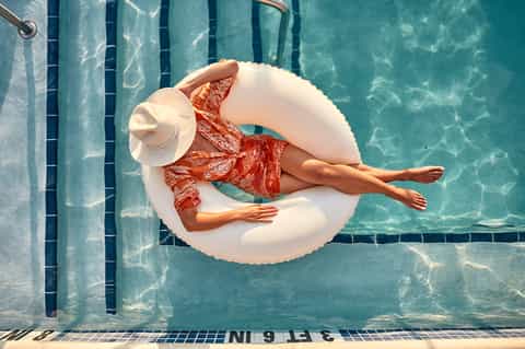 Woman floating in white ring on turquoise pool water wearing red patterned swimsuit and white hat