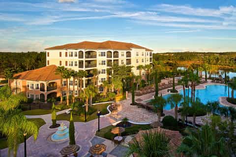 Aerial view of luxury resort with multi-story building, pools, palm trees, and surrounding grounds