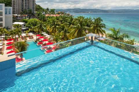 Oceanfront pool with red lounge chairs overlooking turquoise waters and palm trees