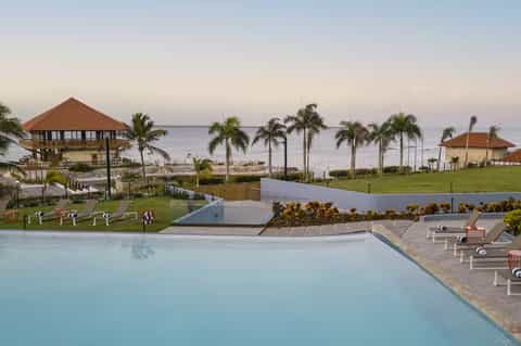 Beachfront resort with multiple blue pools, palm trees, ocean views, and tropical landscape at sunset