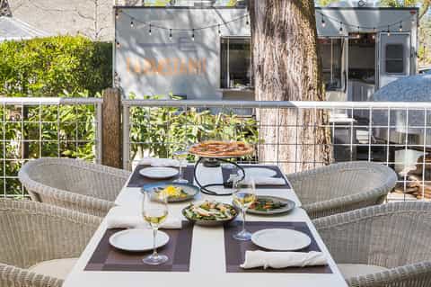 Outdoor dining area with wicker chairs, set table with wine glasses, salad dishes, and farmhouse restaurant in background