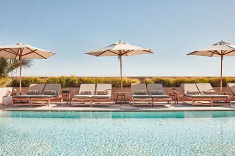 Resort pool area with tan loungers, beige umbrellas, and coastal grassland landscape under clear blue sky