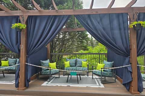 Covered deck with dark blue curtains, wooden beams, and lounge seating with green accent pillows