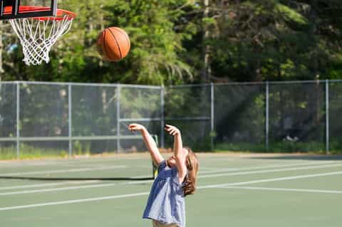 Young girl shooting basketball on outdoor court with green playing surface and chain-link fence