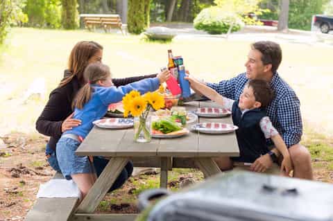 Family of four toasting with drinks at outdoor picnic table with yellow flowers and food
