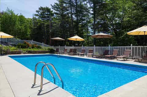 Resort pool with blue water, white railings, lounge chairs, and yellow umbrellas surrounded by pine trees