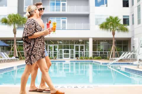 Couple holding tropical cocktails walking by resort pool with palm trees