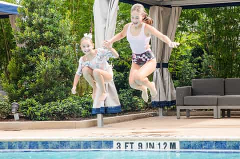 Two children jumping into a shallow pool beside a shaded patio area with lush green landscaping