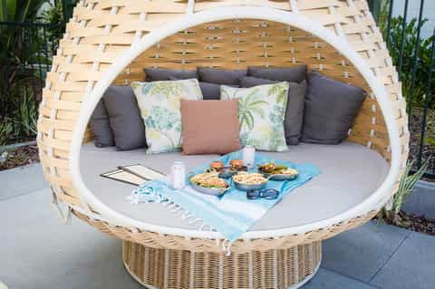 Woven basket daybed with gray cushions, decorative pillows, and food spread on blue picnic cloth