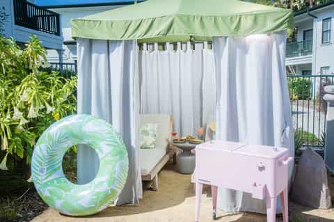 Outdoor cabana tent with green canopy, white curtains, pink cooler, and float ring