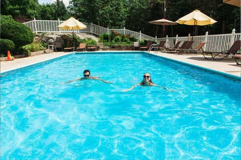 Resort swimming pool with two guests swimming, yellow umbrellas, lounge chairs, and tree-lined landscape