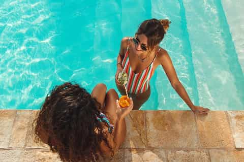 Two women by turquoise pool, one in striped swimsuit handing drink to person in water