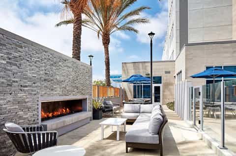 Luxury outdoor patio with fireplace, lounge seating, palm trees, and blue umbrellas