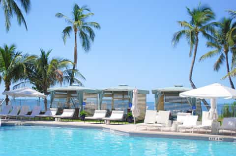 Resort pool with palm trees, white lounge chairs, wooden cabanas, and ocean view under clear blue sky