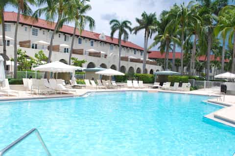 Resort pool with turquoise water, white umbrellas, lounge chairs, palm trees, and Spanish colonial-style building with red tile roof