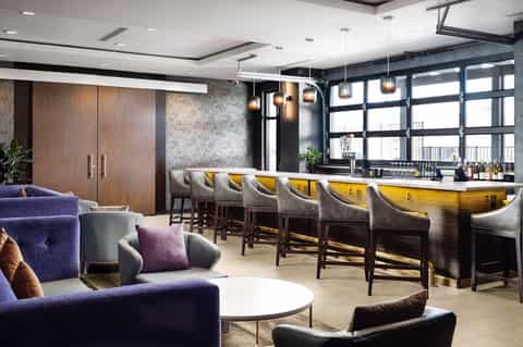 Modern hotel bar and lounge with purple seating, yellow-lit bar counter, and pendant lighting