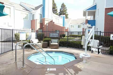 Outdoor spa hot tub surrounded by black fencing with residential buildings and pine trees in background