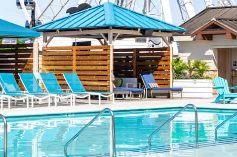 Resort pool area with turquoise lounge chairs, wooden pergola with blue roof, sailboats visible in background