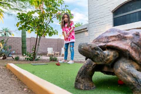 Young girl playing mini golf with turtle statue obstacle in desert resort courtyard with palm trees
