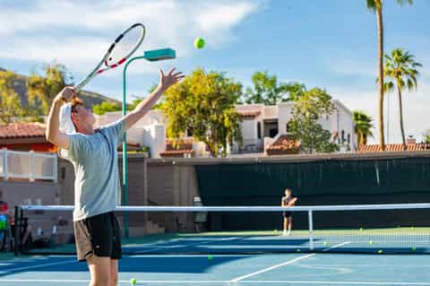 Man serving tennis ball on outdoor court with palm trees and residential buildings in background