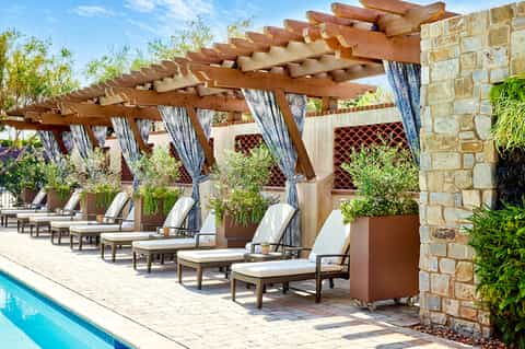 Poolside cabanas with wooden pergola and blue curtains, stone walls, and lounge chairs