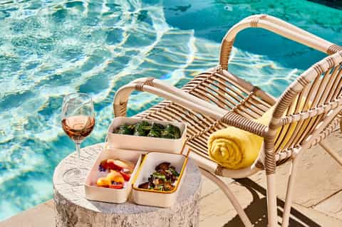 Poolside lounge chair with rose wine, gourmet food containers, and yellow towel by sparkling water