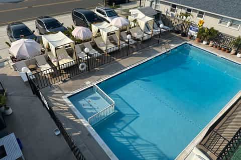 Resort pool deck with blue swimming pool, white umbrellas, lounge chairs, and parked cars in background