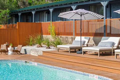 Resort pool deck with wooden loungers, striped umbrella, modern house with orange railing, and lush landscaping