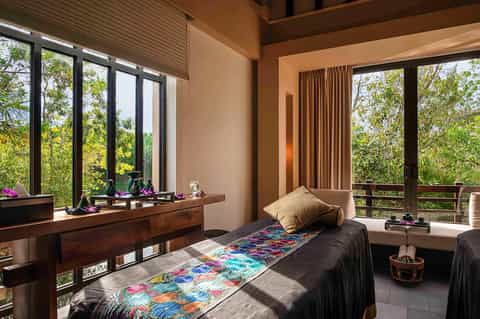 Modern bedroom with black bed, colorful throw blanket, wooden desk, and floor-to-ceiling windows overlooking tropical forest