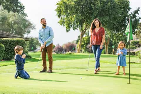 Family playing miniature golf on a manicured green lawn with trees and a flag in background