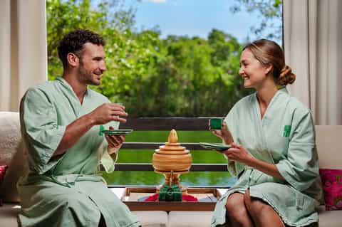 Couple in spa robes enjoying tea and conversation with lush green views through windows