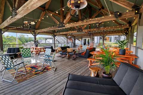 Covered outdoor deck with wooden beams, string lights, colorful lounge chairs, and orange dining tables overlooking green landscape