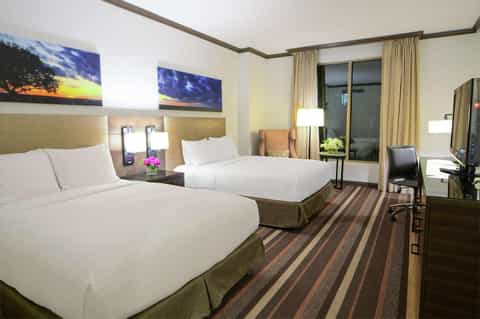 Hotel room with two queen beds, tan headboards, artwork, desk area, and balcony with curtains
