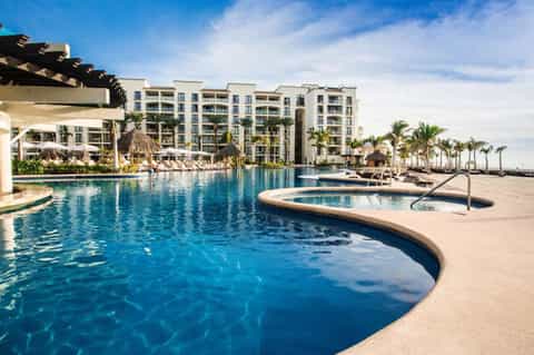 Beachfront resort pool complex with lounge chairs, palm trees, and multi-story hotel buildings under blue sky