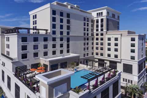 Modern multi-story white hotel complex with rooftop pool, lounge areas, and blue swimming pool surrounded by purple flowering plants