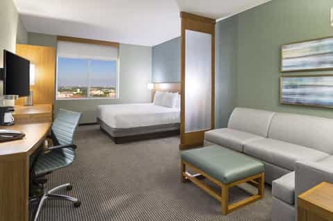 Modern hotel suite with king bed, sofa, work desk, flat-screen TV, and cityscape views through floor-to-ceiling windows