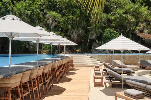 Resort pool deck with woven bar seating, white umbrellas, and loungers surrounded by tropical vegetation