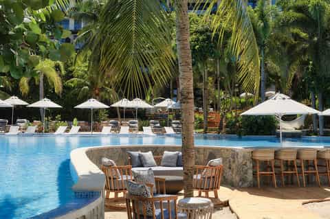 Tropical resort pool with loungers, umbrellas, and palm trees surrounding curved seating area in water