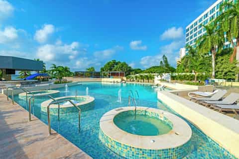 Resort pool complex with multiple hot tubs, palm trees, loungers, and modern building under blue sky