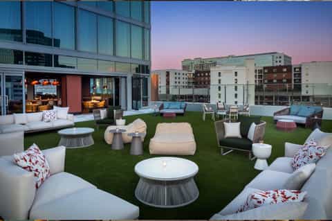 Rooftop lounge with artificial turf, modern seating clusters, fire pits, and interior space overlooking city skyline at dusk