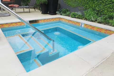 Small residential swimming pool with brick border, built-in steps, and metal handrail surrounded by concrete patio